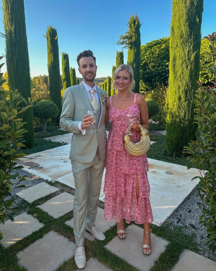 A man in a grey suit and a woman in a pink floral dress posing in a garden.