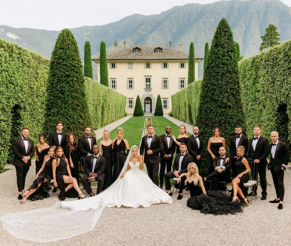A wedding party in black formal wear posing before a large villa and mountains.