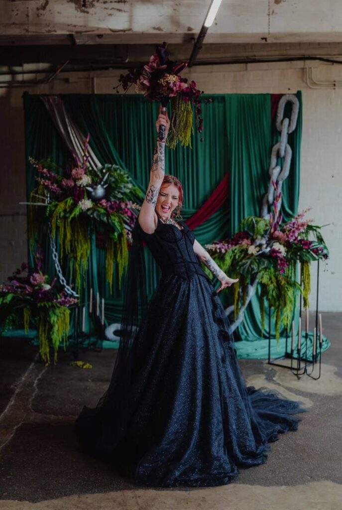 A woman in a sparkling black ballgown wedding dress poses with a bouquet against a green backdrop.