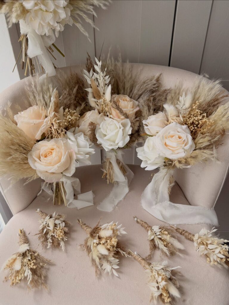 Three cream rose and dried pampas grass bouquets with ribbons resting on a pink chair.