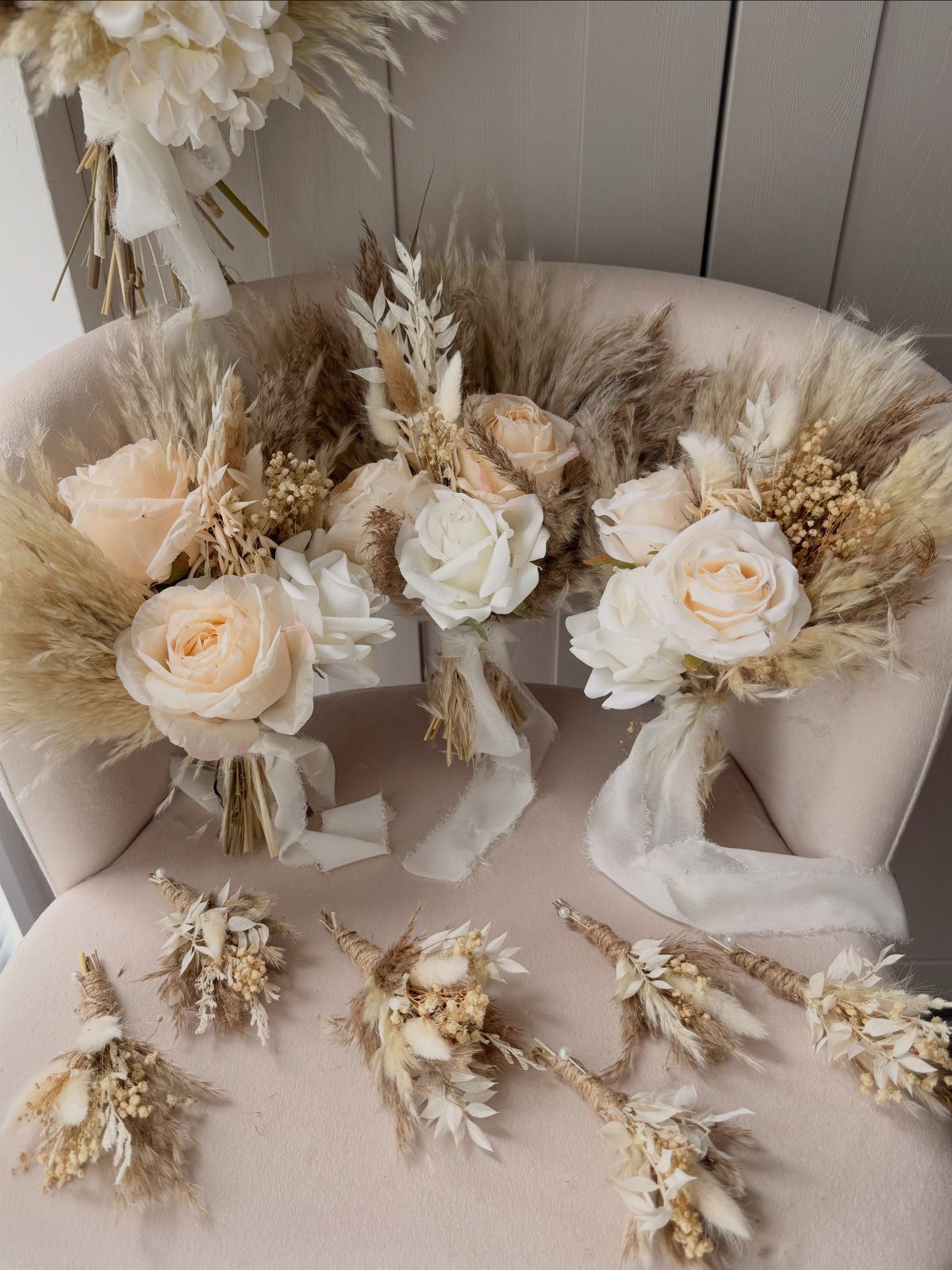 Three cream rose and dried pampas grass bouquets with ribbons resting on a pink chair.