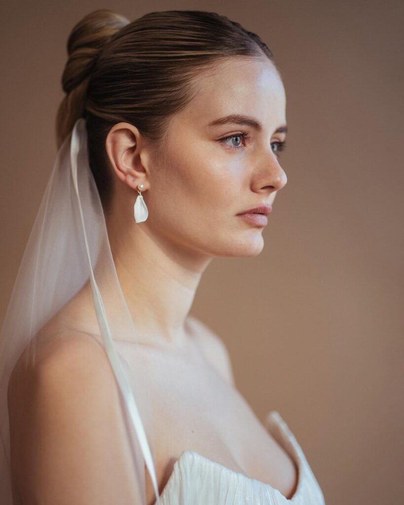 A woman with a sleek blonde updo wearing a white bridal veil and drop earrings.