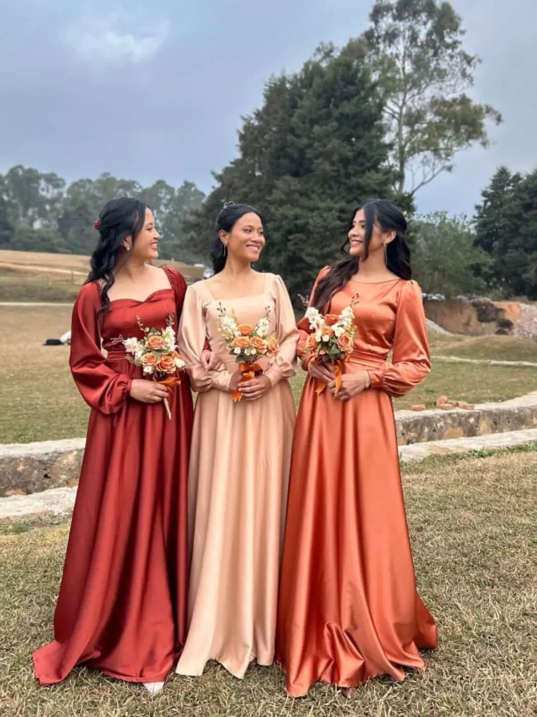 Three bridesmaids in long satin autumn-colored dresses holding floral bouquets in a grassy field.