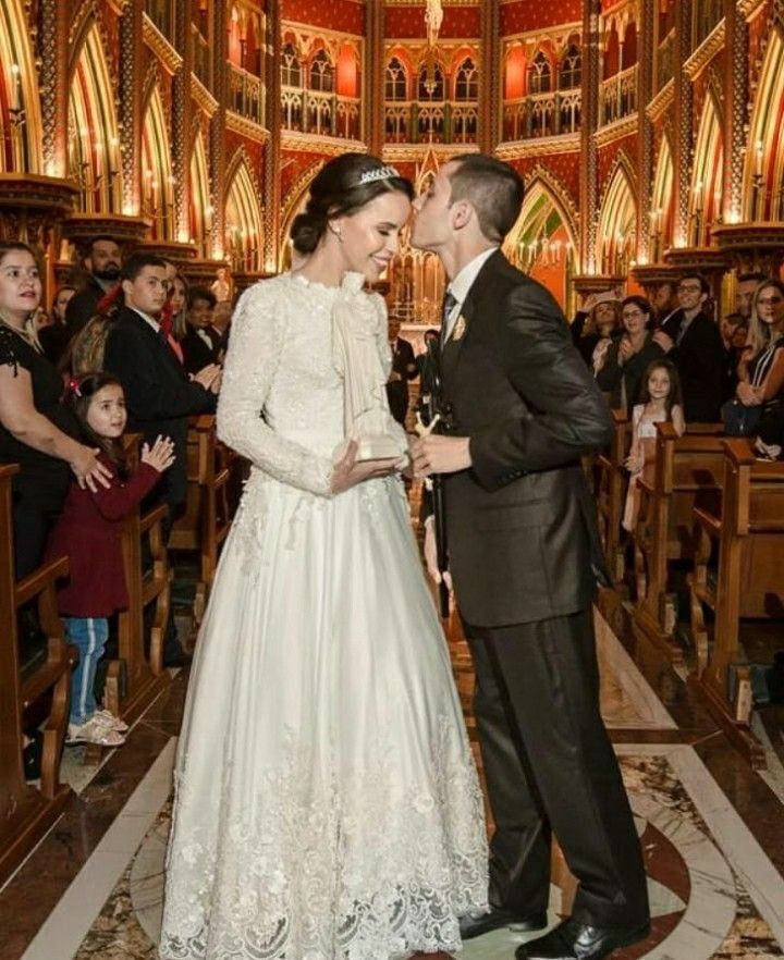 A bride wearing a long-sleeved lace gown kisses her groom in a church aisle.