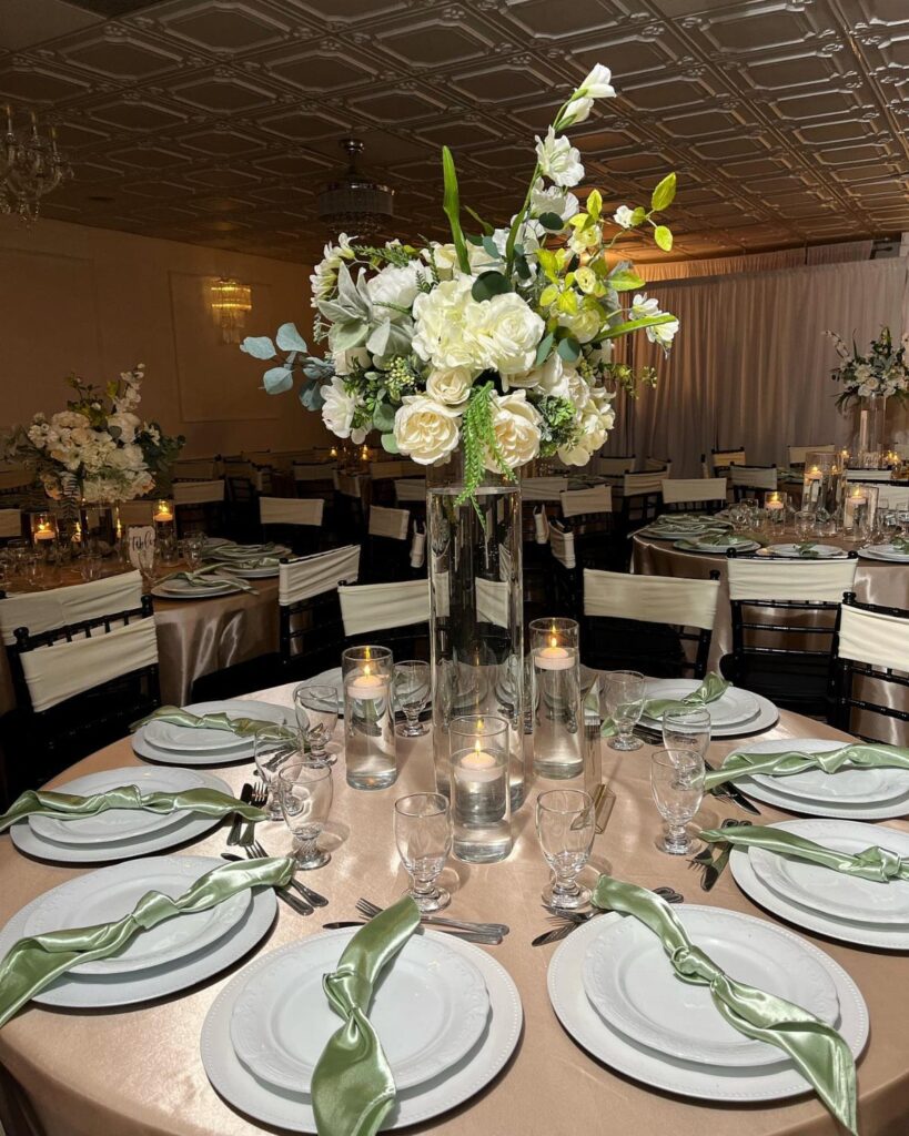 A tall glass vase filled with white roses and sage green eucalyptus greenery on a wedding table.