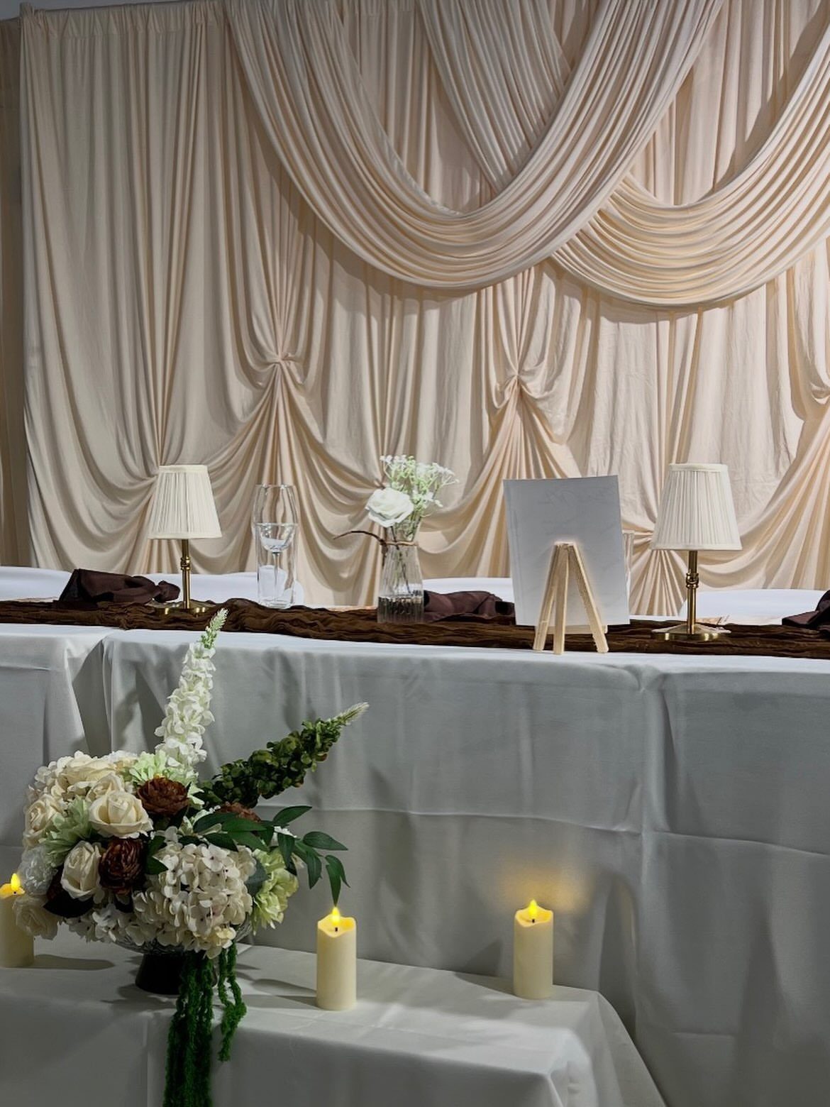 Decorated event table with champagne drapes, white flowers, and lit candles against a white tablecloth.