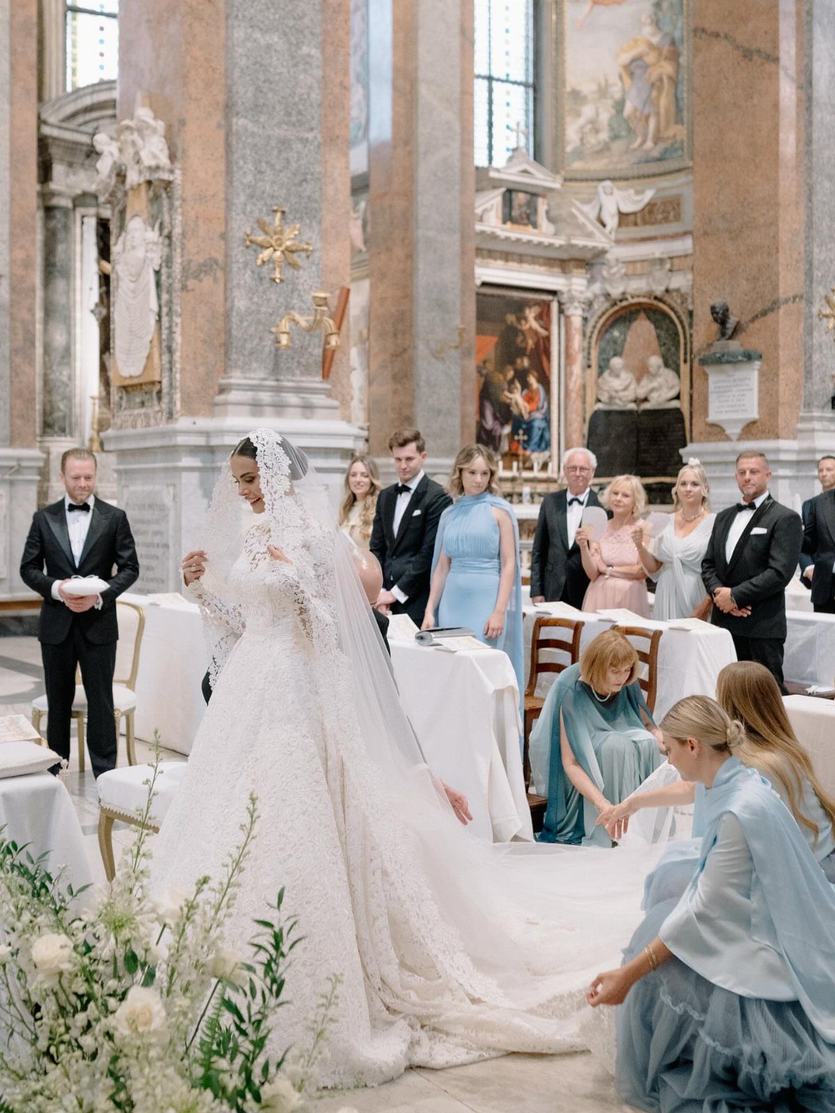 A bride in a lace wedding dress and veil stands inside a grand, ornate church.