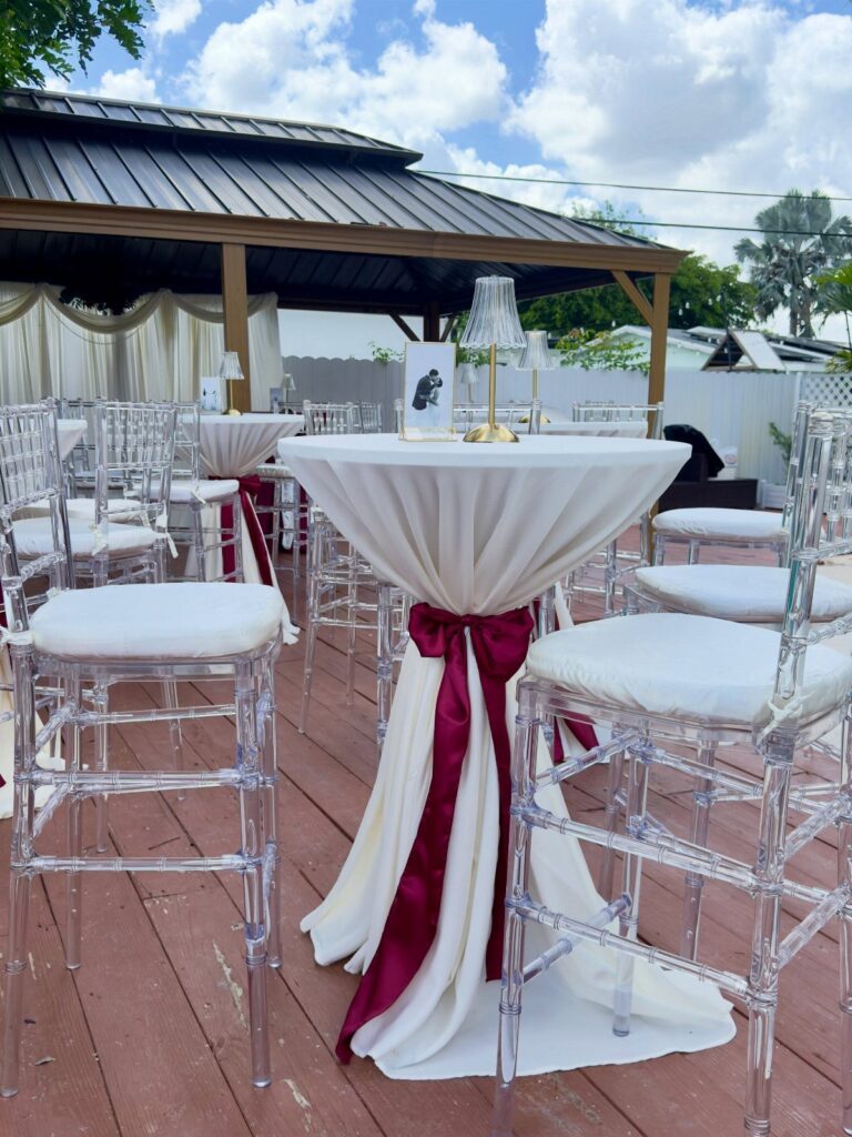 A white draped cocktail table with a burgundy ribbon and a small lamp on a wooden deck.