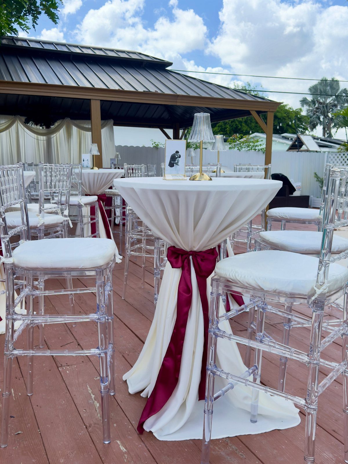A white draped cocktail table with a burgundy ribbon and a small lamp on a wooden deck.