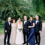 A wedding party posing together in formal attire amidst lush green trees in a park.