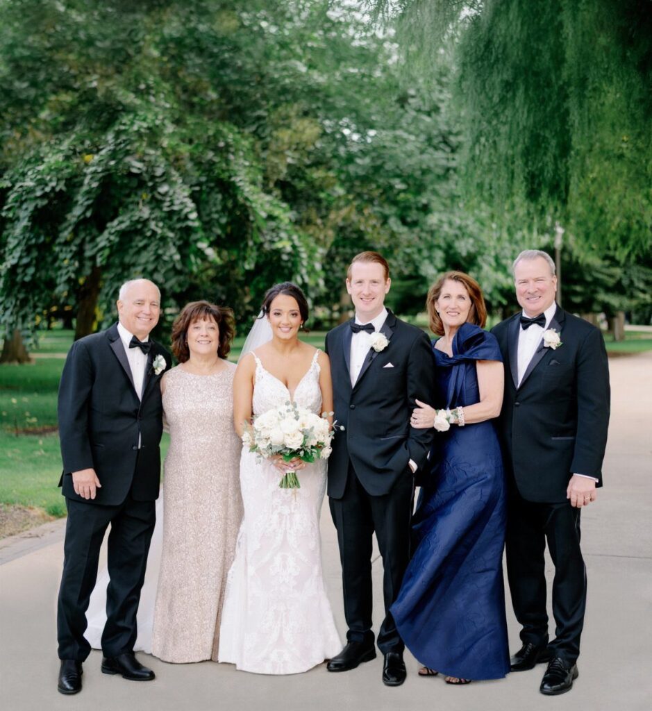 A wedding party posing together in formal attire amidst lush green trees in a park.