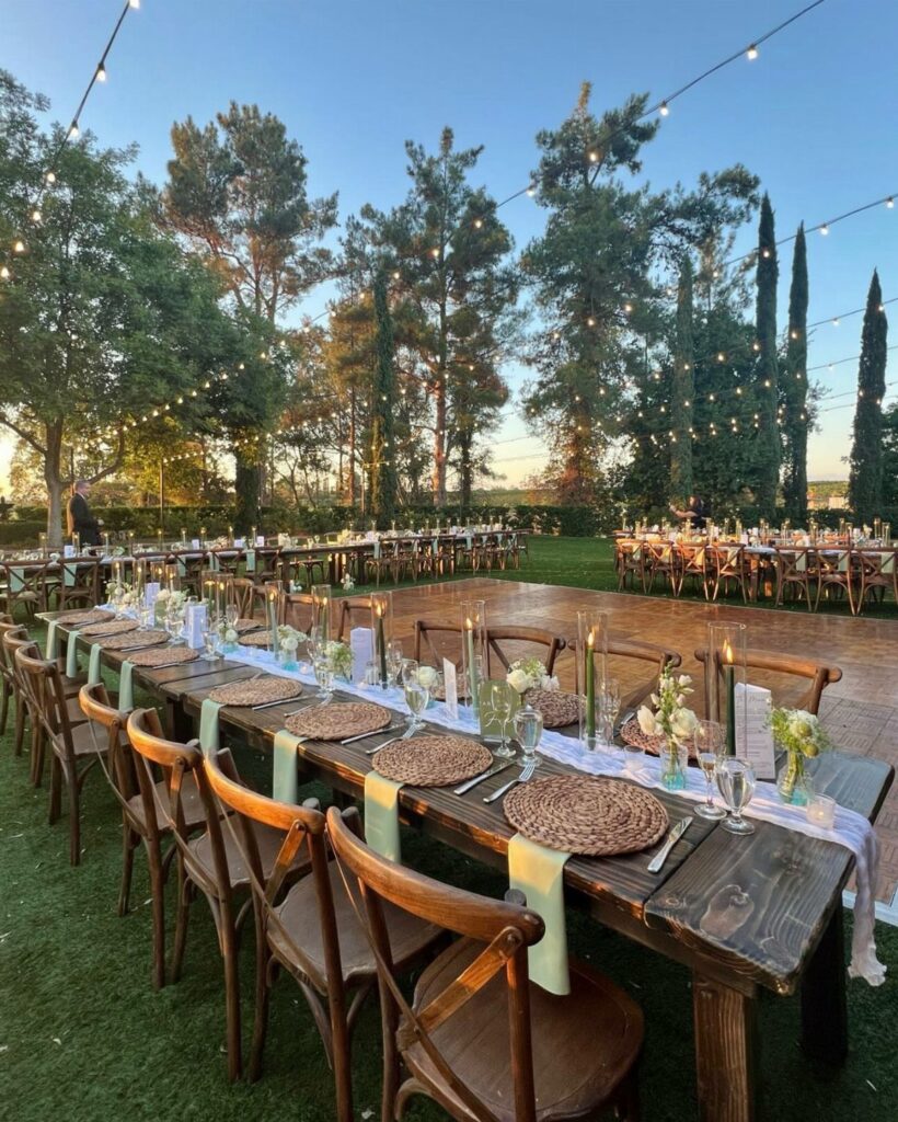 Long wooden farm tables set with white runners and greenery for an outdoor wedding reception.