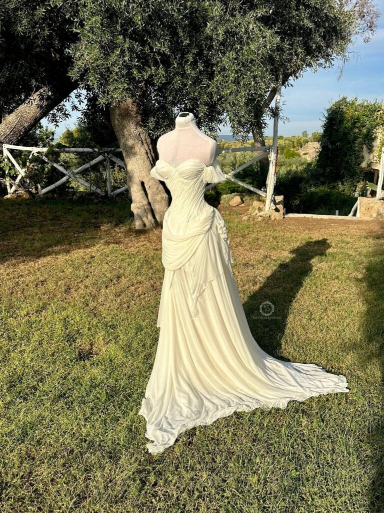 A white off-the-shoulder wedding dress with a long train displayed on a mannequin outdoors.