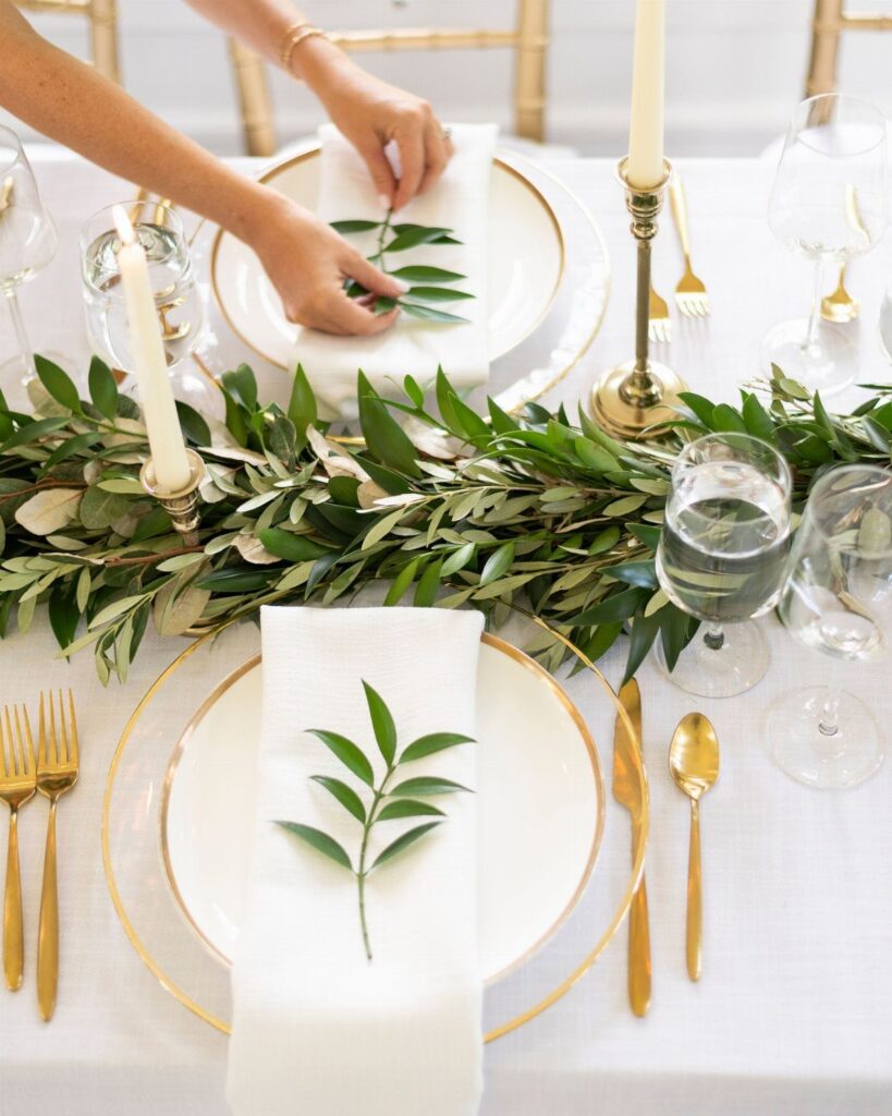 Greenery garland and gold tableware set on a white tablecloth with hands arranging a napkin sprig.