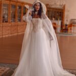 A woman in a white wedding dress with a long, sheer hooded cape standing in a ballroom.