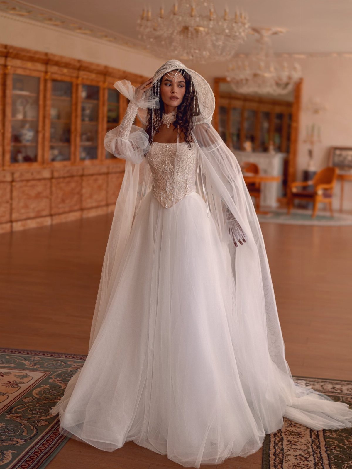 A woman in a white wedding dress with a long, sheer hooded cape standing in a ballroom.