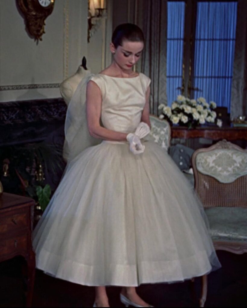 A woman in a white sleeveless tea-length gown and gloves standing in an elegant room.