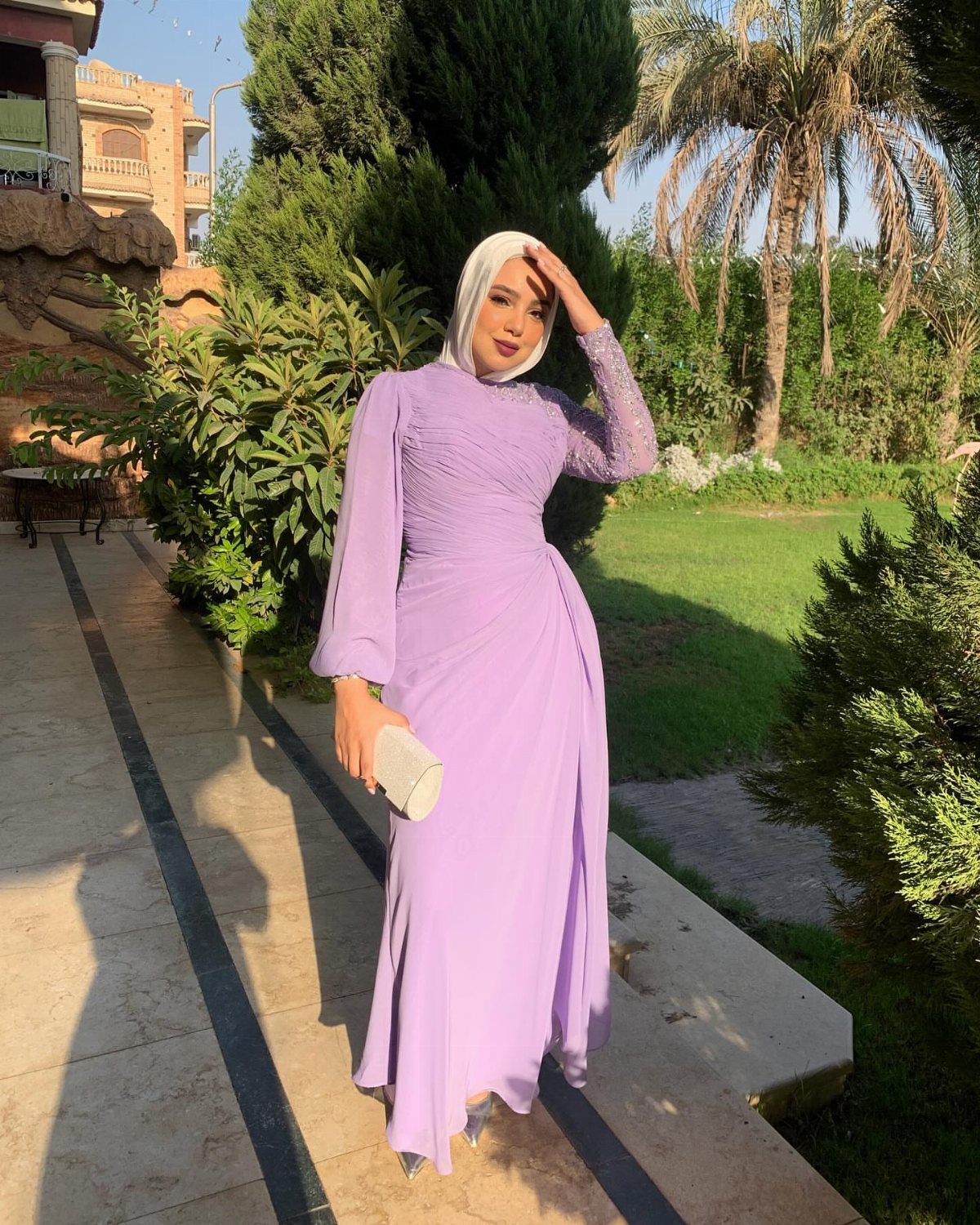 A woman in a long lavender dress and white hijab posing outdoors near greenery.