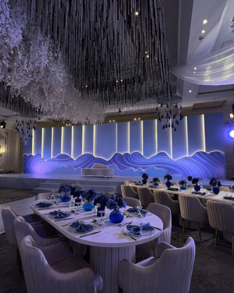 Wedding reception tables set with royal blue napkins and glassware under a silver fringe ceiling installation.