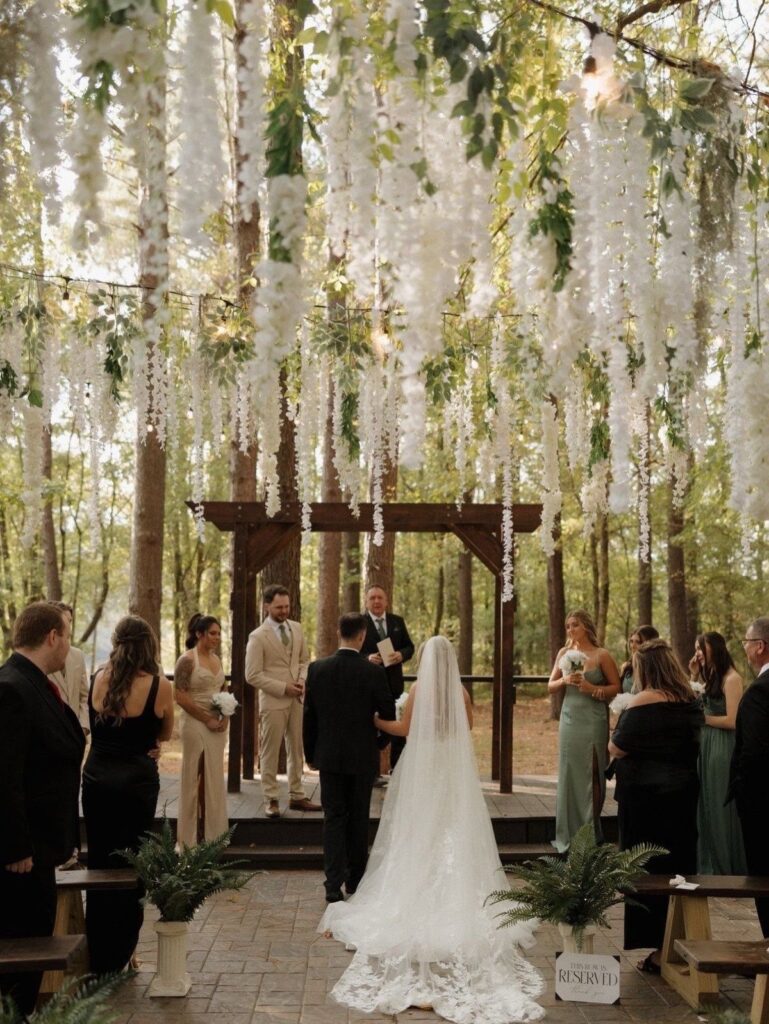 A bride and groom stand under a wooden arch decorated with hanging white flowers in a forest.