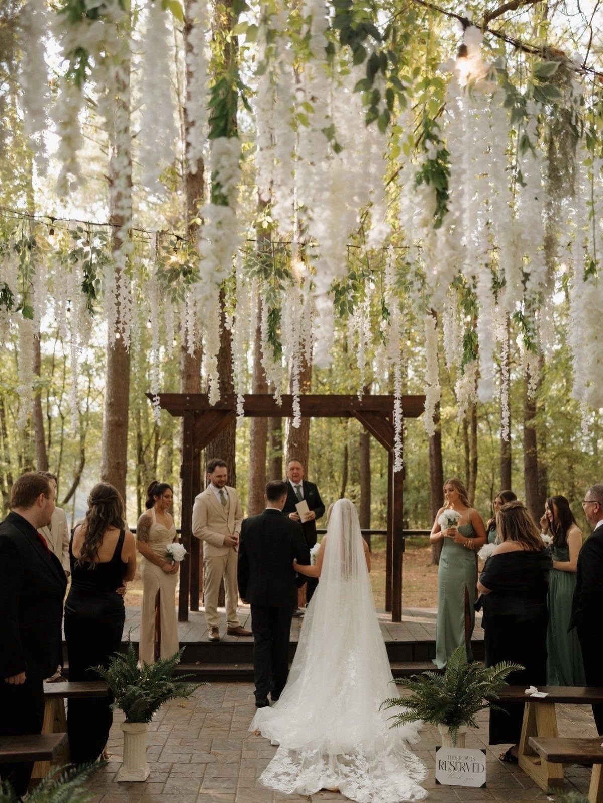A bride and groom stand under a wooden arch decorated with hanging white flowers in a forest.