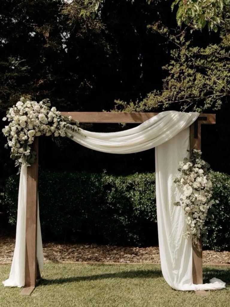 A wooden wedding arbor draped in white fabric and adorned with white floral arrangements outdoors.