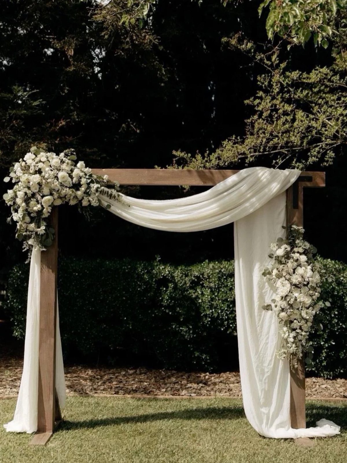 A wooden wedding arbor draped in white fabric and adorned with white floral arrangements outdoors.