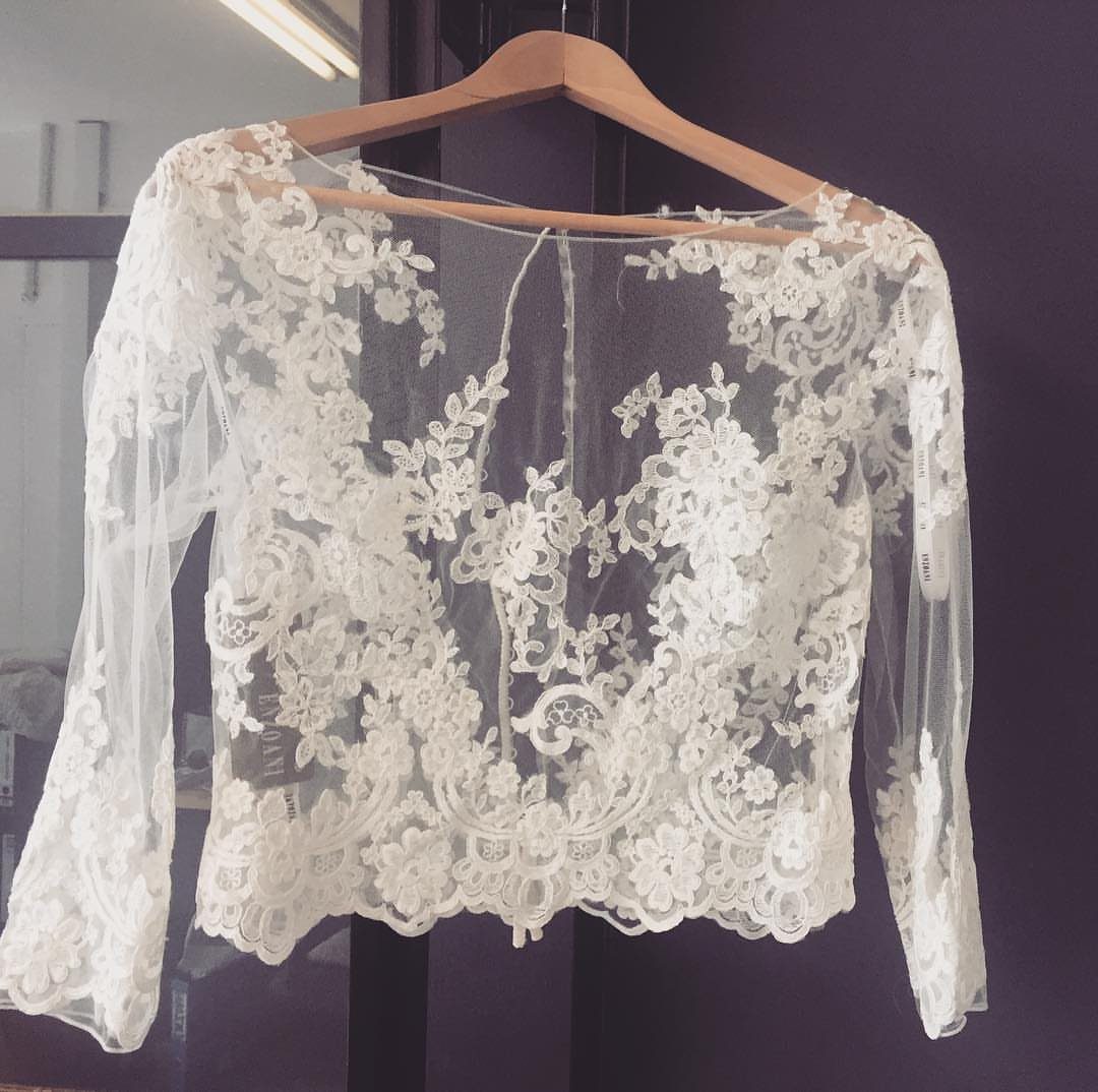 A white lace bolero jacket with floral patterns hanging on a wooden hanger.