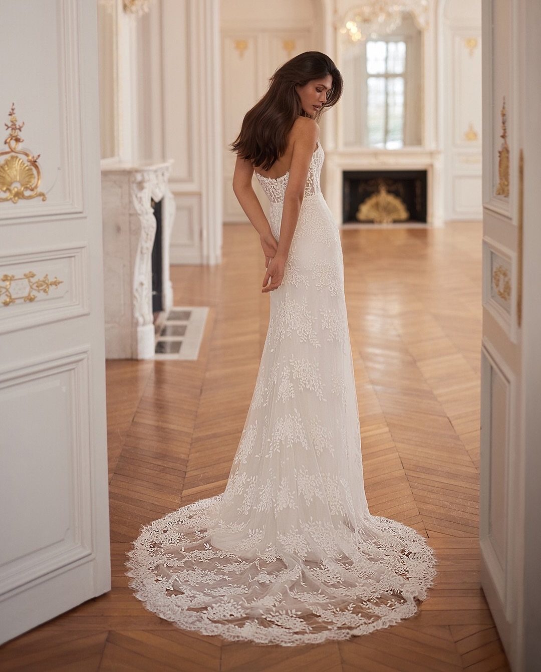 A woman in a white lace wedding dress with a long train standing in an ornate room.
