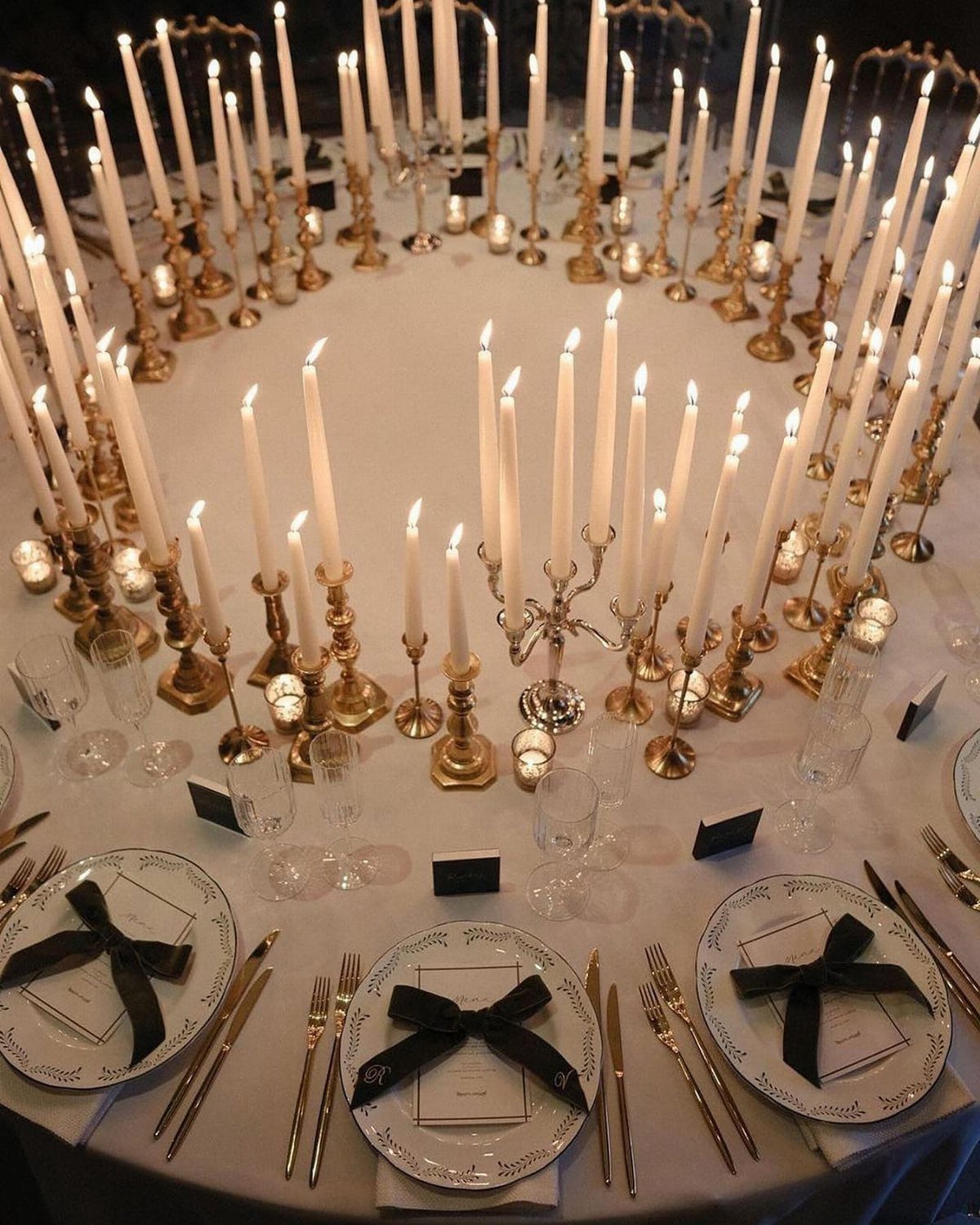 Formal wedding table setting with white plates, black bows, gold cutlery, and many lit taper candles.