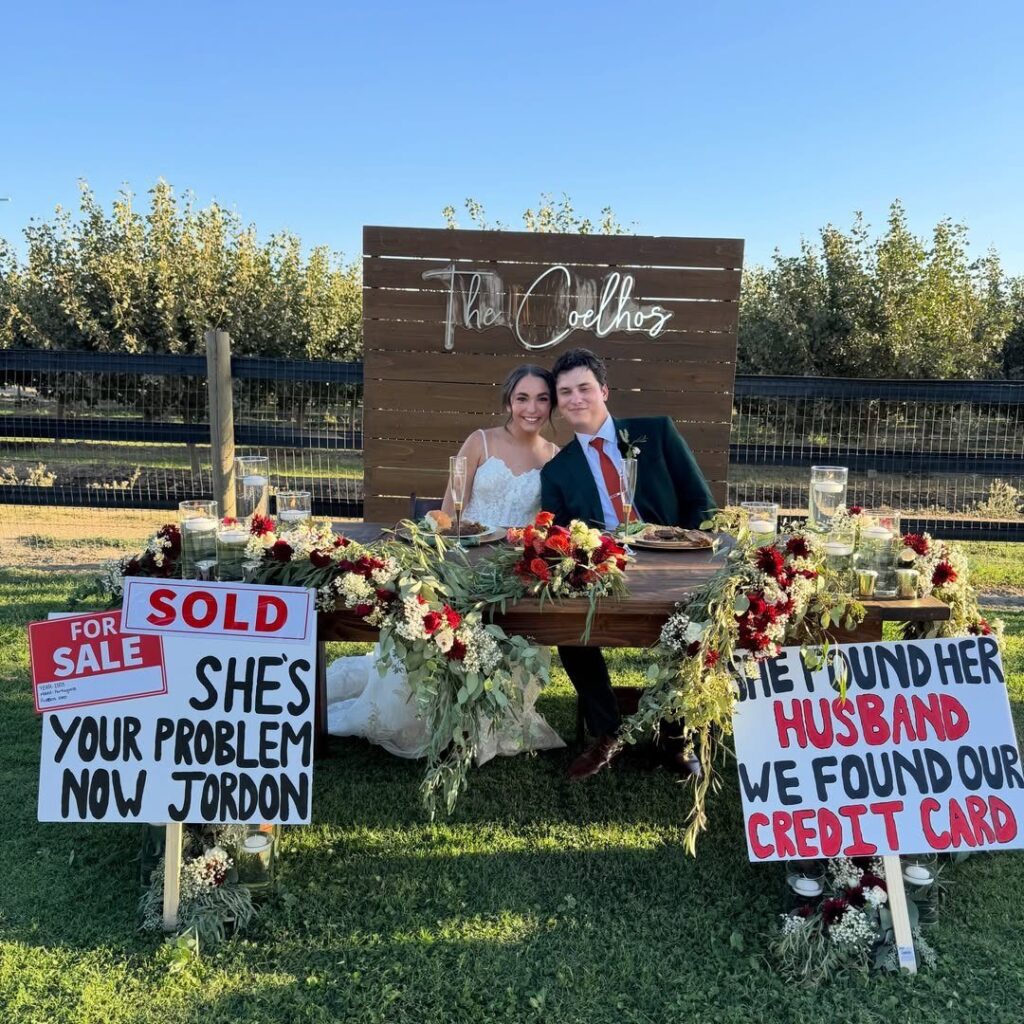 A bride and groom sit behind funny wedding signs on a decorated outdoor table.