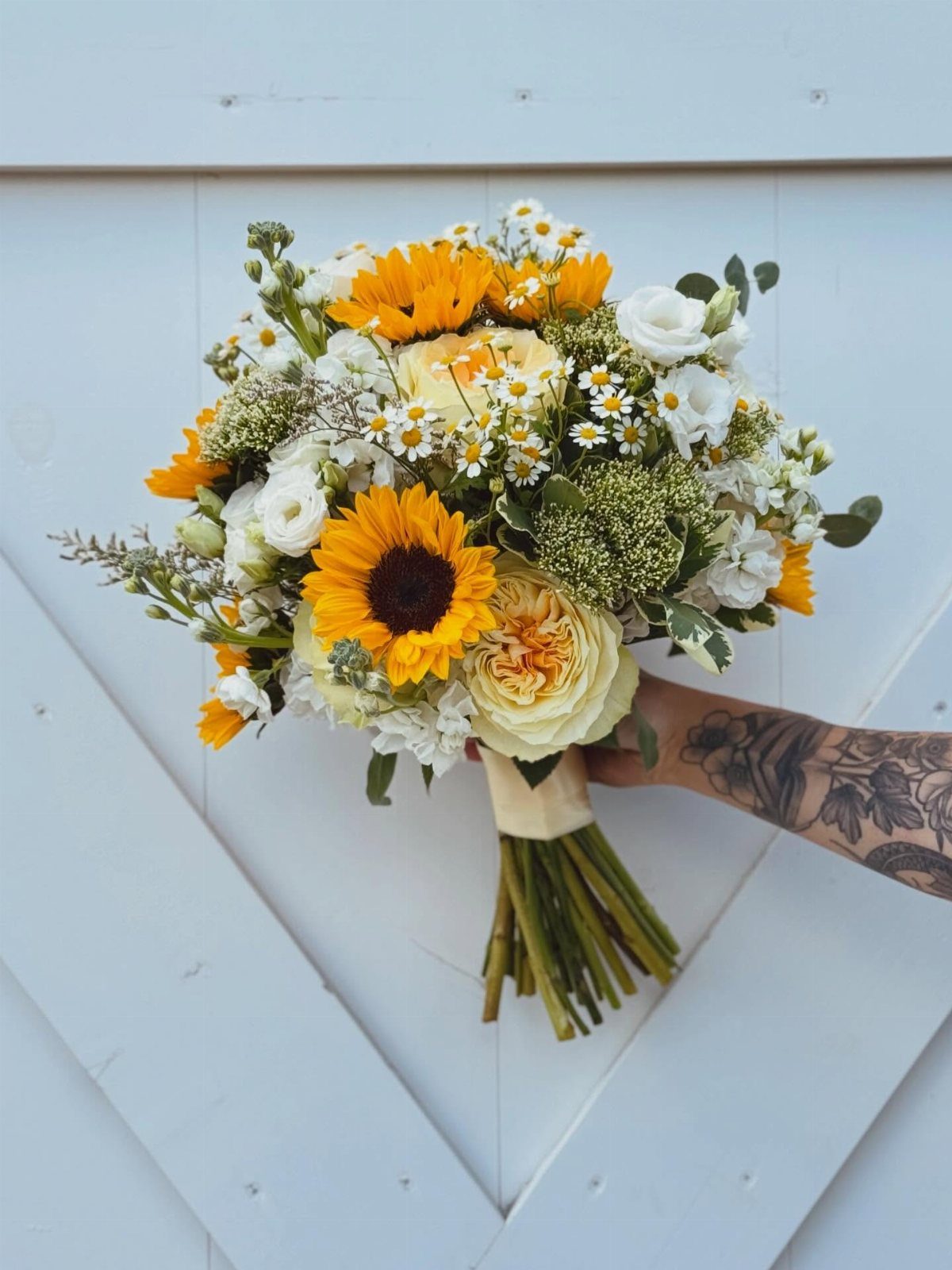 A wedding bouquet with sunflowers, white roses, and daisies held against a white wooden wall.