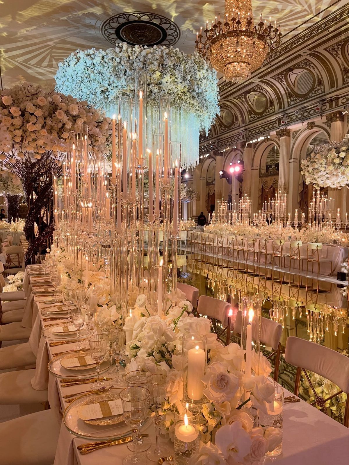White floral centerpieces and tall candles decorate long banquet tables in a grand ballroom.