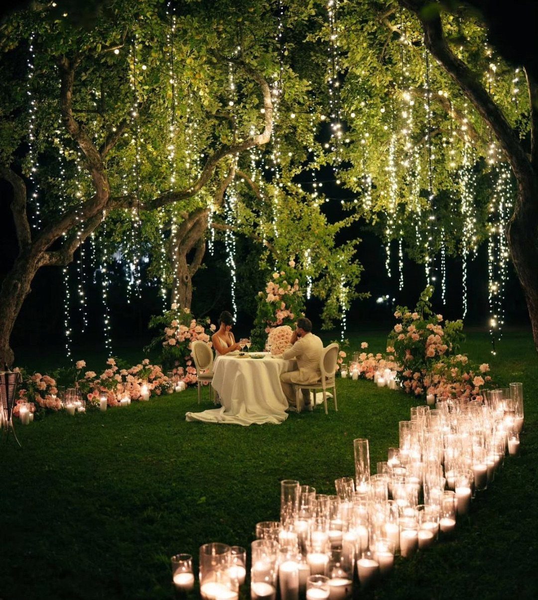 A couple dines under a large tree decorated with hanging vertical fairy lights and surrounded by candles.