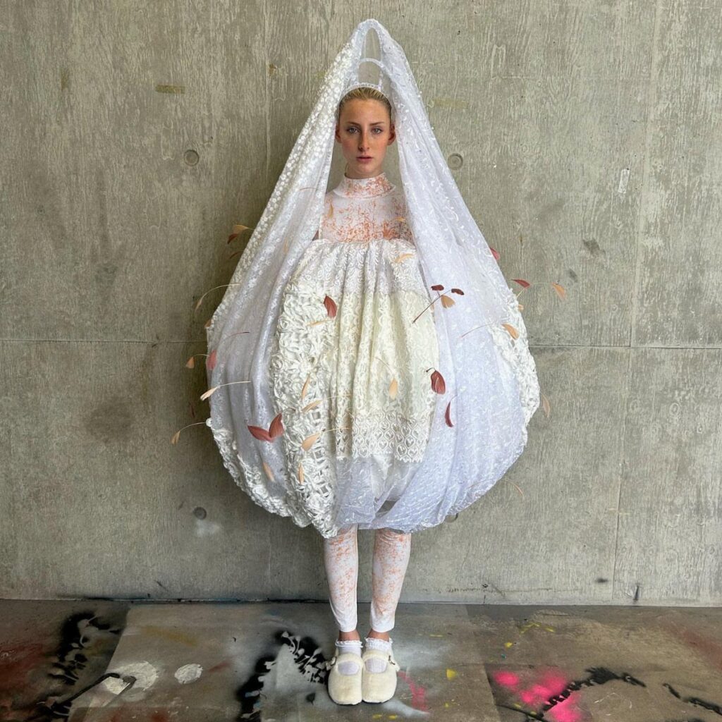 A woman wearing a voluminous, avant-garde white lace wedding dress and tall veil.