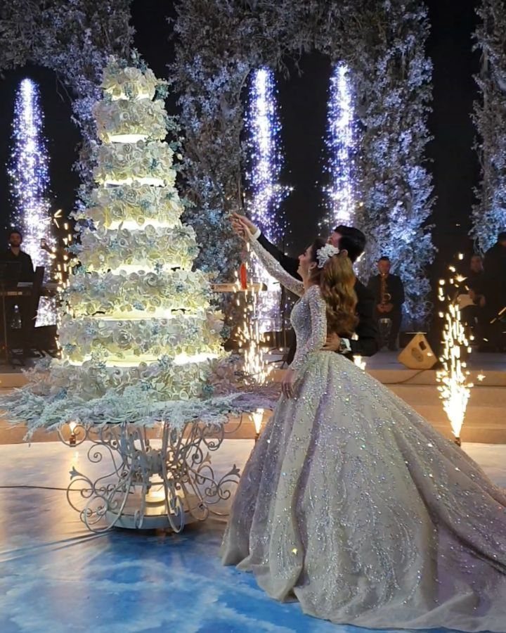 A bride in a shimmering gown stands beside a towering white floral wedding cake under winter lights.